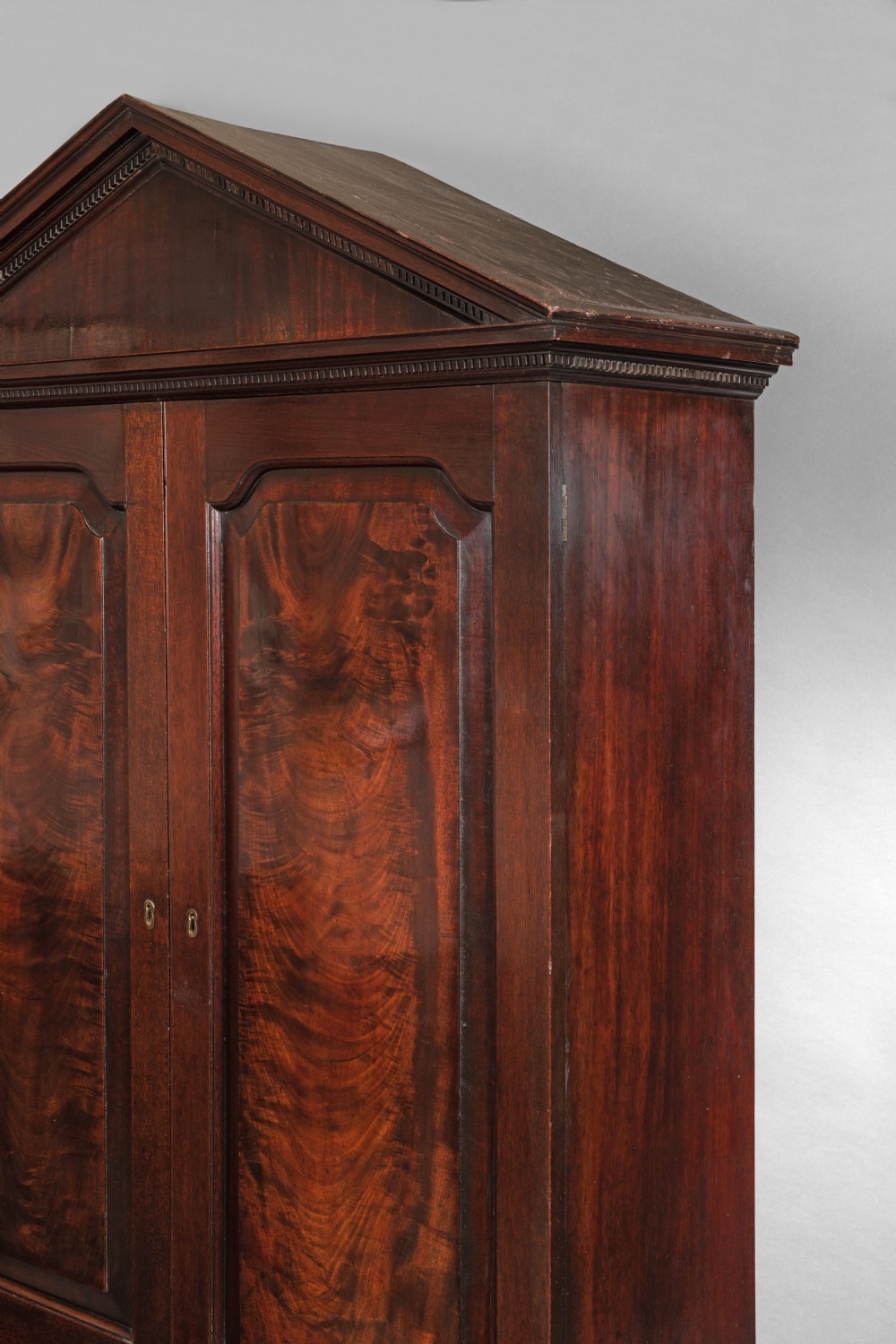 An antique Georgian mahogany linen press featuring a pediment top, two panelled doors with flame mahogany veneer, three drawers below, and bracket feet. View II