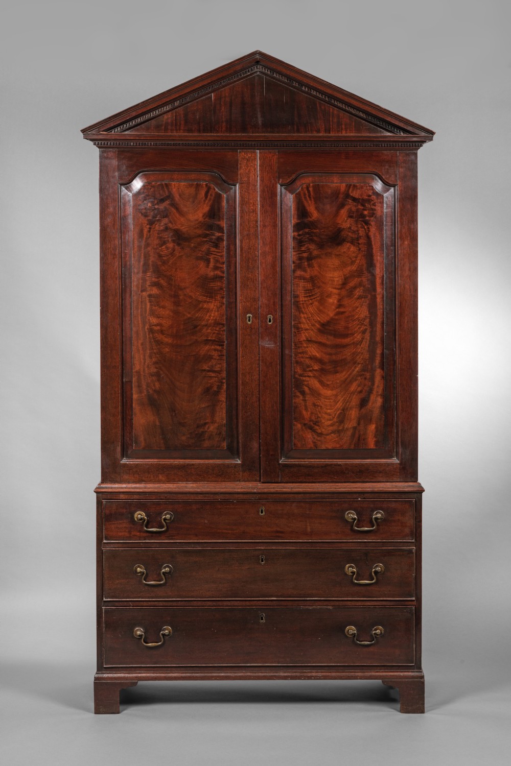 An antique Georgian mahogany linen press featuring a pediment top, two panelled doors with flame mahogany veneer, three drawers below, and bracket feet.