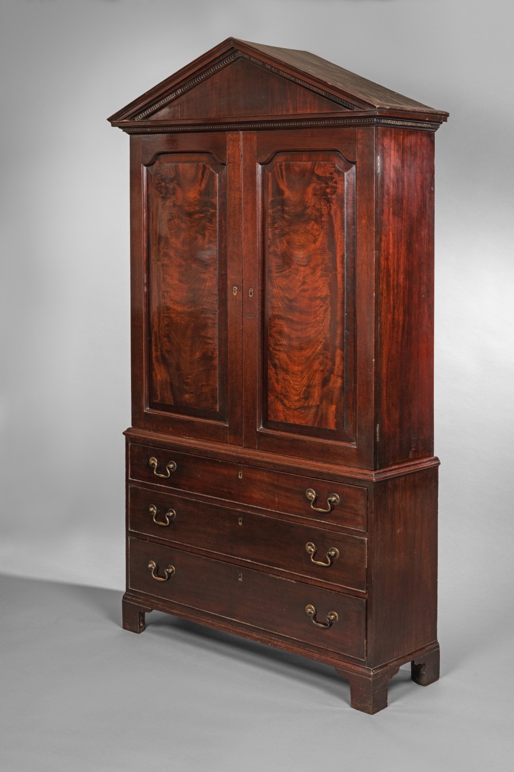 An antique Georgian mahogany linen press featuring a pediment top, two panelled doors with flame mahogany veneer, three drawers below, and bracket feet. View IV