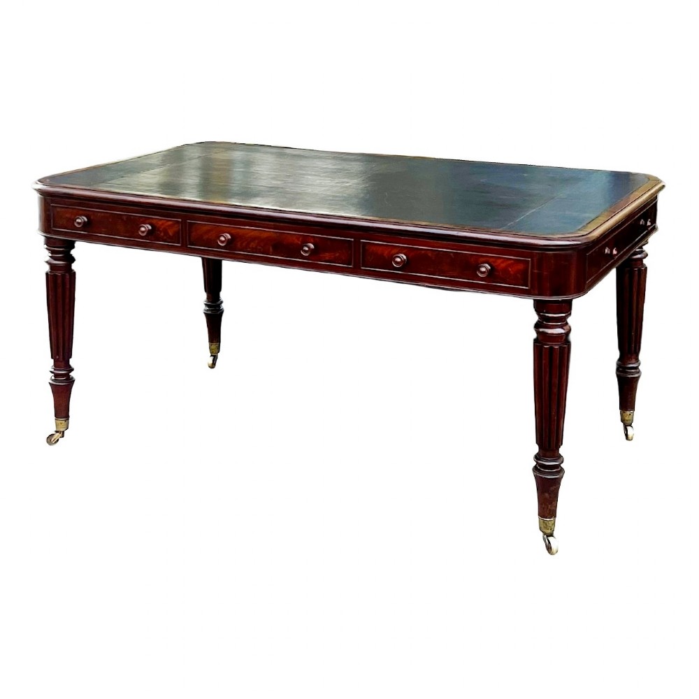 A Regency mahogany writing table featuring a dark leather insert top, three frieze drawers with round wooden knobs, and reeded tapering legs with brass castors.