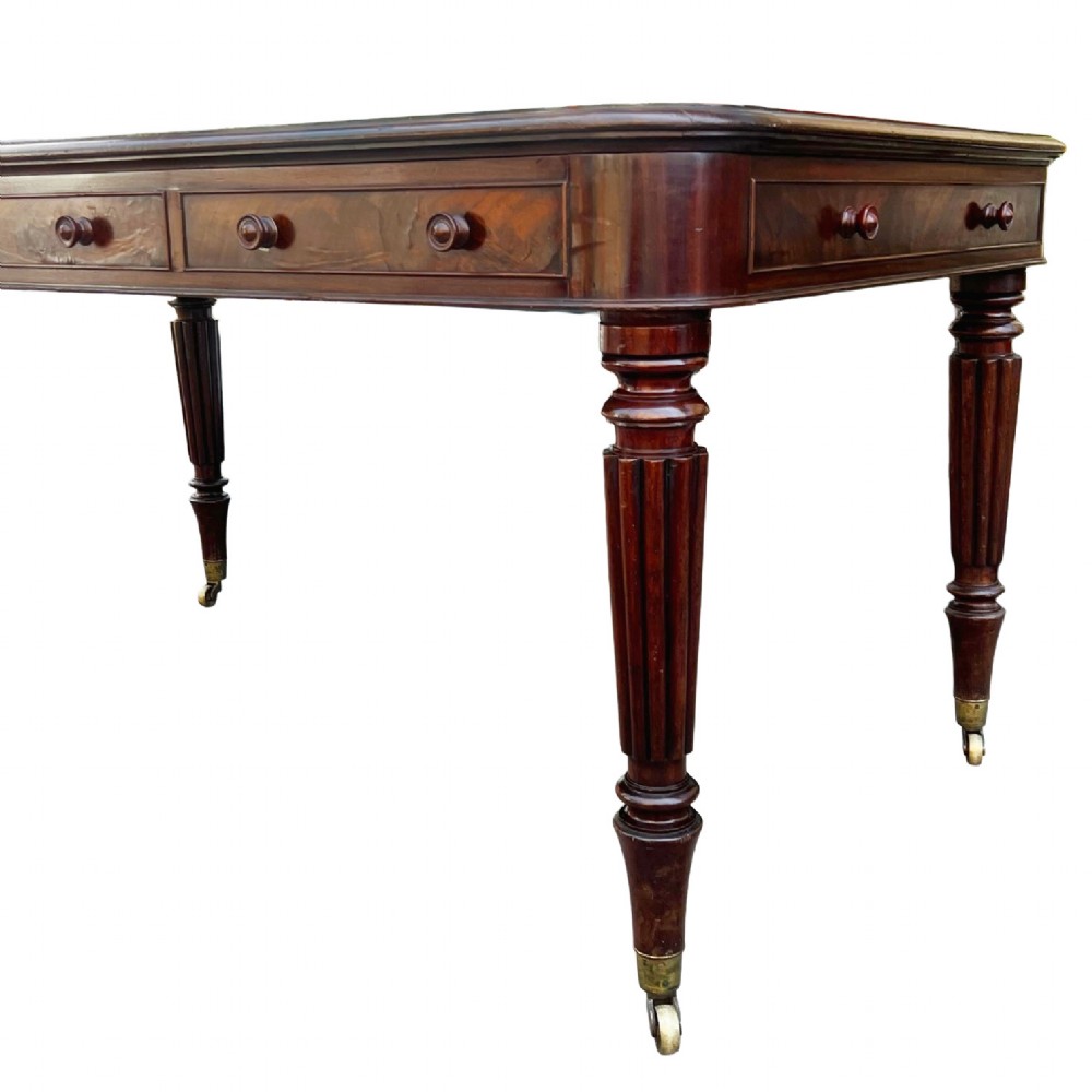 A Regency mahogany writing table featuring a dark leather insert top, three frieze drawers with round wooden knobs, and reeded tapering legs with brass castors. View II