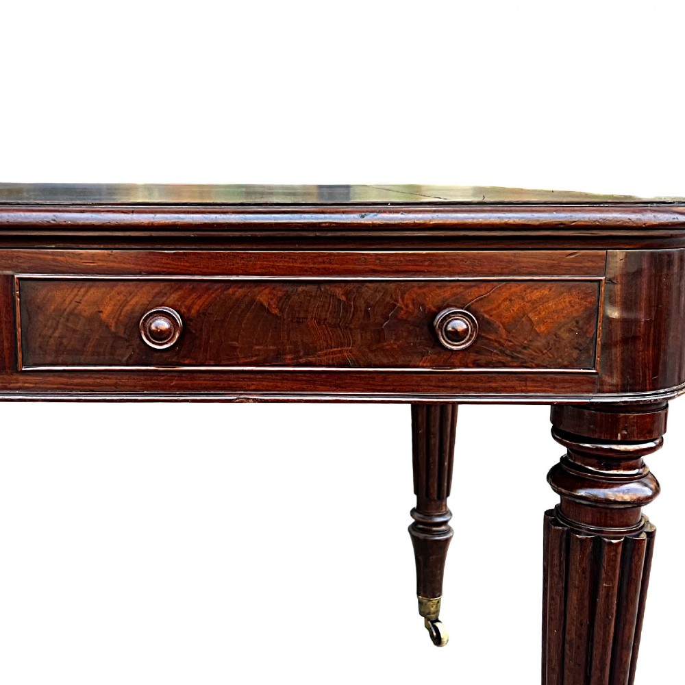 A Regency mahogany writing table featuring a dark leather insert top, three frieze drawers with round wooden knobs, and reeded tapering legs with brass castors. View III