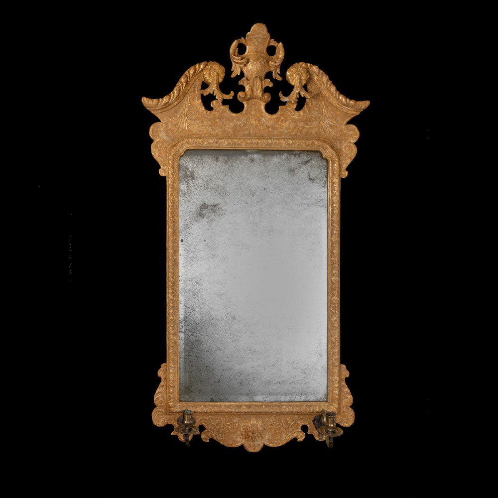 An eighteenth-century George II carved gilt gesso wall mirror featuring an architectural pediment with cresting, low-relief floral patterns, foxed mirror glass, and two brass candle sconces at the base.
