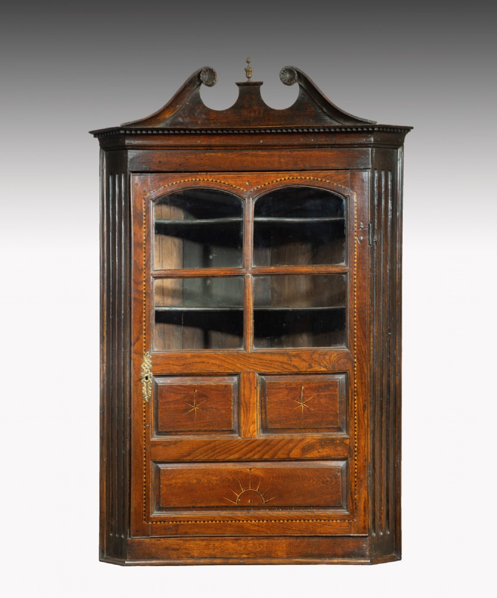 An 18th-century Georgian dark oak corner cupboard featuring a swan-neck pediment, glazed upper door panels, and decorative inlay motifs.