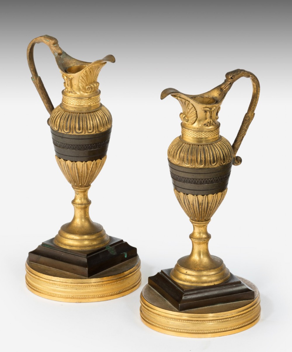 A pair of 19th-century Neoclassical ewers featuring reeded gilt-bronze urn bodies with dark patinated bronze central bands, cast handles, and tiered bases.