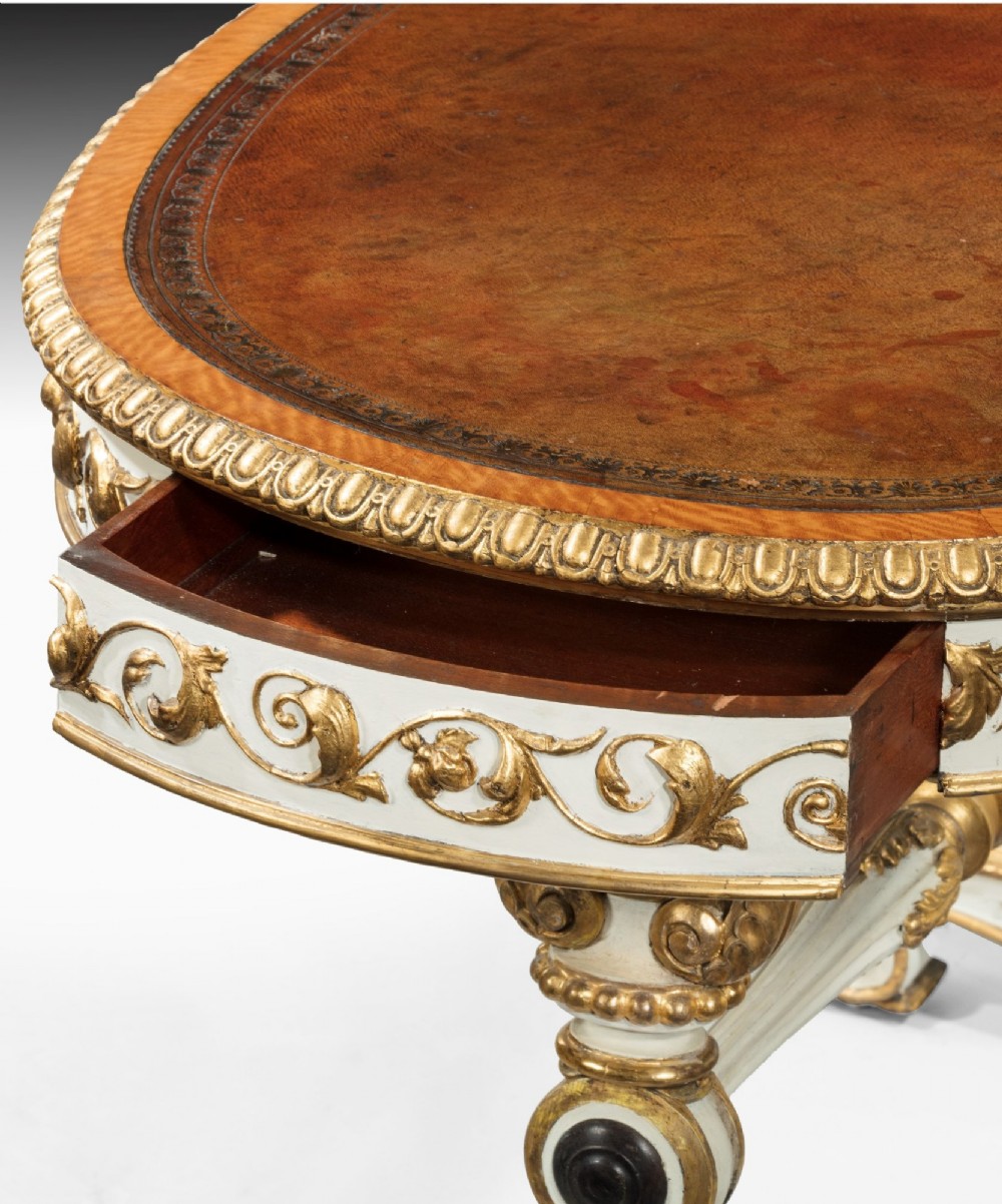 A kidney-shaped William IV writing table featuring an ivory-painted finish, detailed carved giltwood embellishments, a turned stretcher, and a brown leather inset surface. View IV