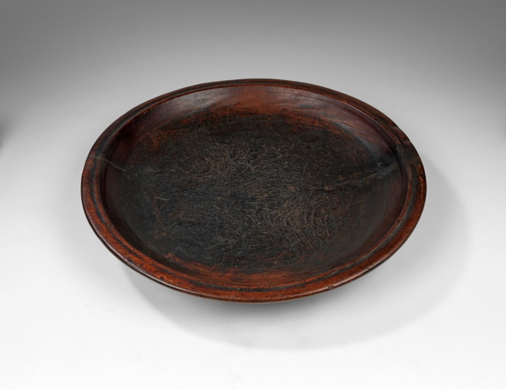 A circular, hand-turned dark wooden bowl with a shallow basin, defined outer rim, and aged grain patina. View II