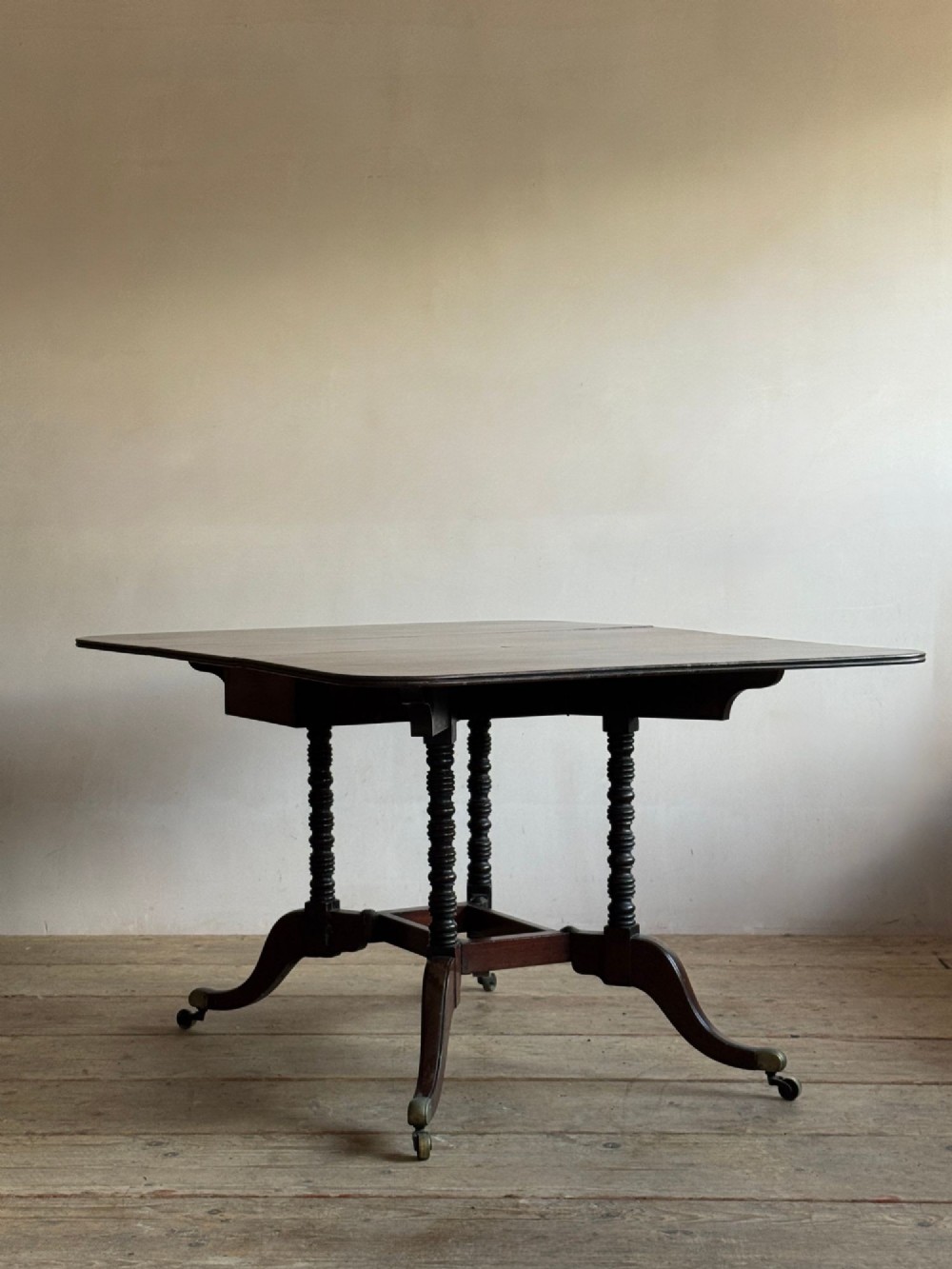 A dark mahogany Georgian Cumberland drop-leaf dining table with bobbin-turned dual legs, splayed feet, and brass castors.