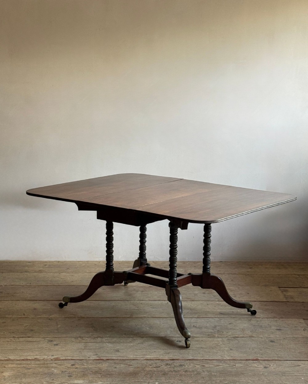 A dark mahogany Georgian Cumberland drop-leaf dining table with bobbin-turned dual legs, splayed feet, and brass castors. View IX