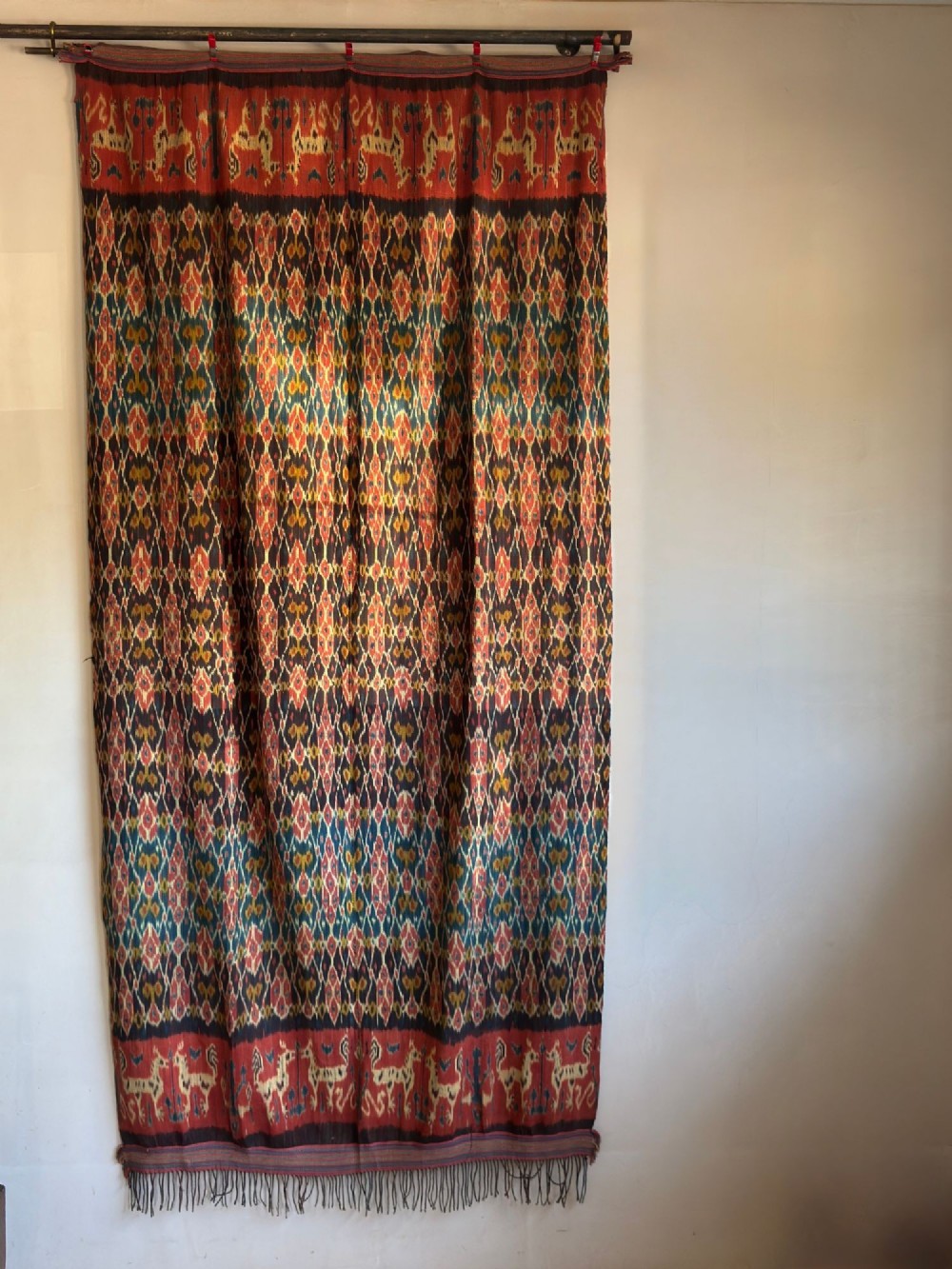 A vertical handwoven Sumba ikat hinggi textile featuring diamond lattice patterns in terracotta red, dark indigo, and yellow with zoomorphic border bands and lower fringe.