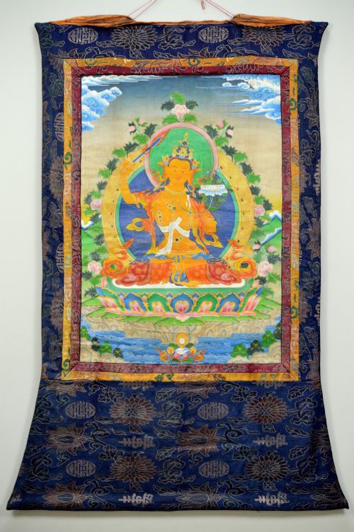 A Tibetan Thangka scroll depicting the seated deity Manjushri holding a raised sword and lotus stem, framed with a patterned silk border. View II
