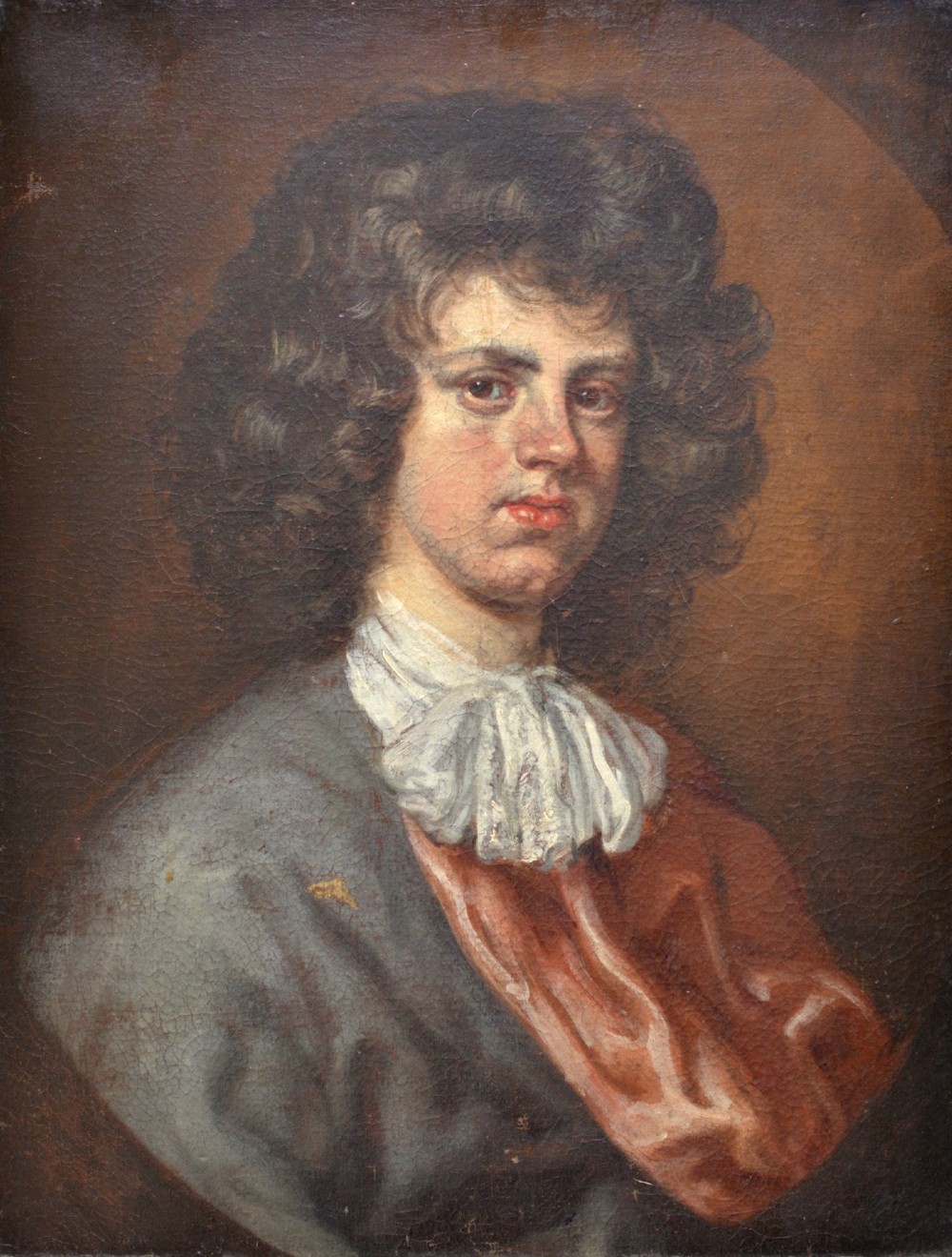 An antique oil portrait painting depicting a young gentleman with dark voluminous curls, wearing a white lace cravat and red mantle within a feigned oval frame.