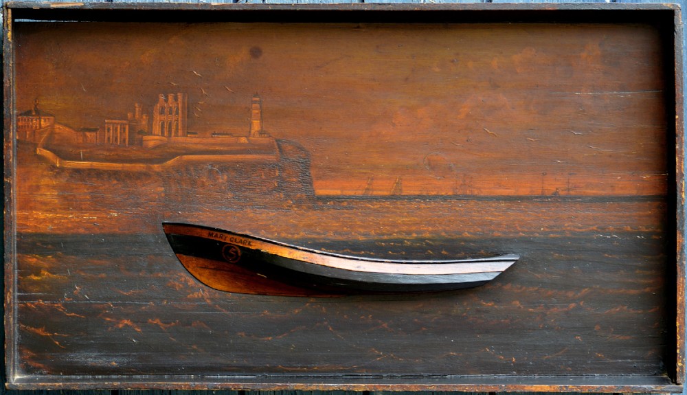 A 19th-century framed wooden folk art diorama featuring a relief-carved half hull coble boat set against a painted view of Tynemouth Castle and Priory.