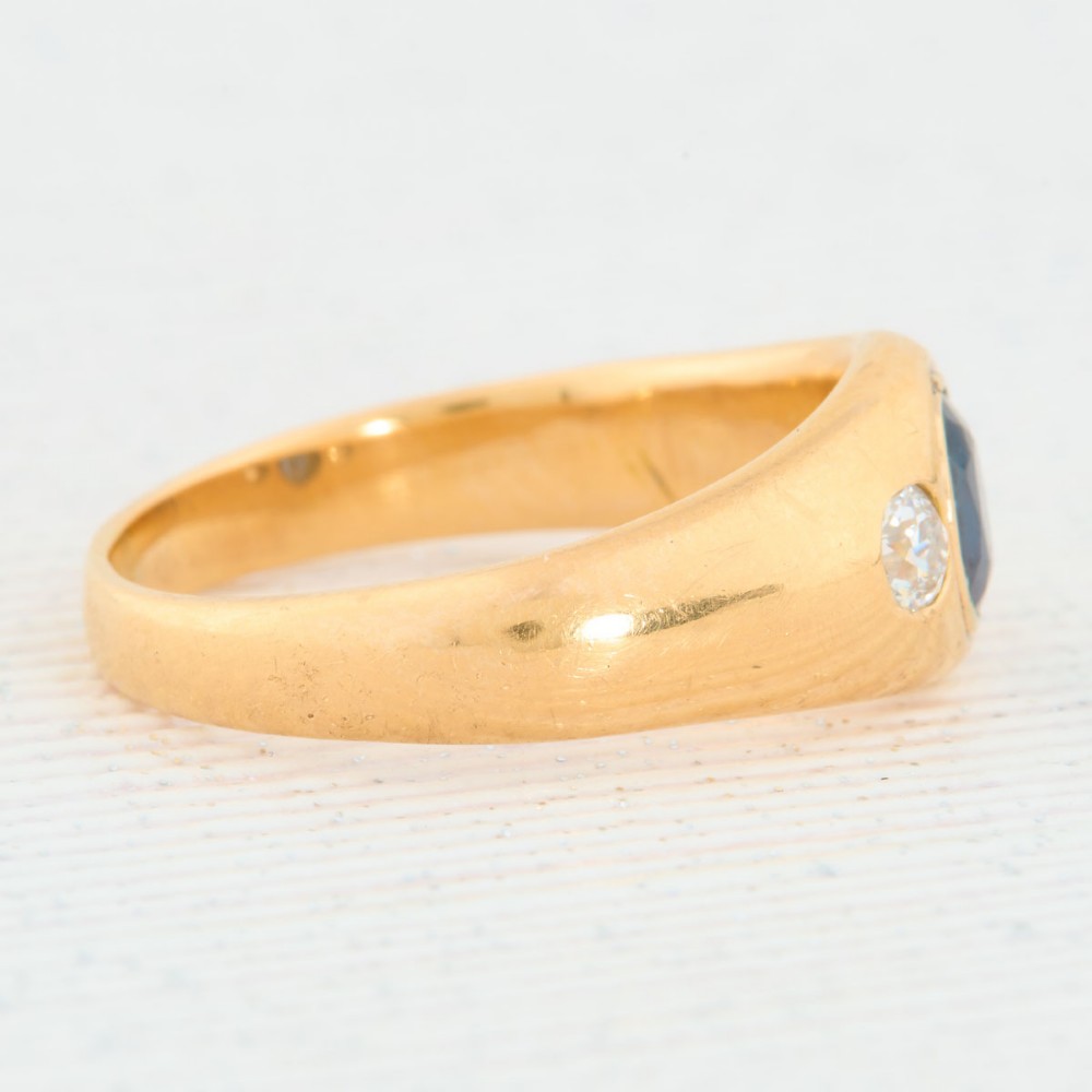 An 18ct yellow gold band features a central oval blue sapphire flanked by two flush-set round diamonds. View IV