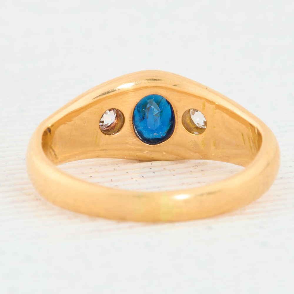 An 18ct yellow gold band features a central oval blue sapphire flanked by two flush-set round diamonds. View V