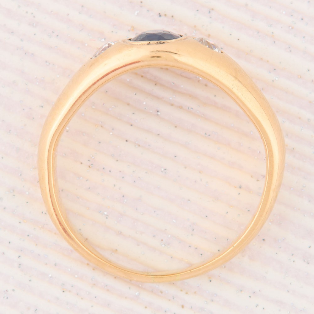 An 18ct yellow gold band features a central oval blue sapphire flanked by two flush-set round diamonds. View VI