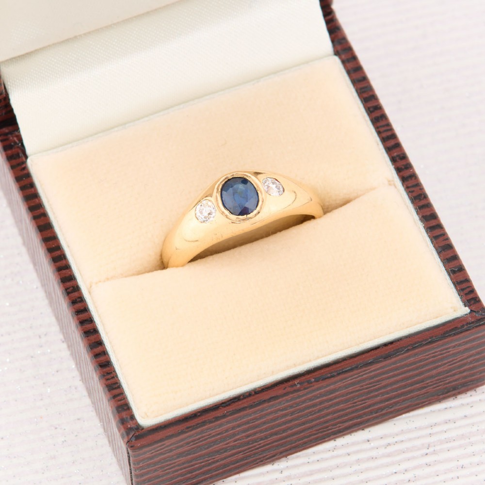 An 18ct yellow gold band features a central oval blue sapphire flanked by two flush-set round diamonds. View VII