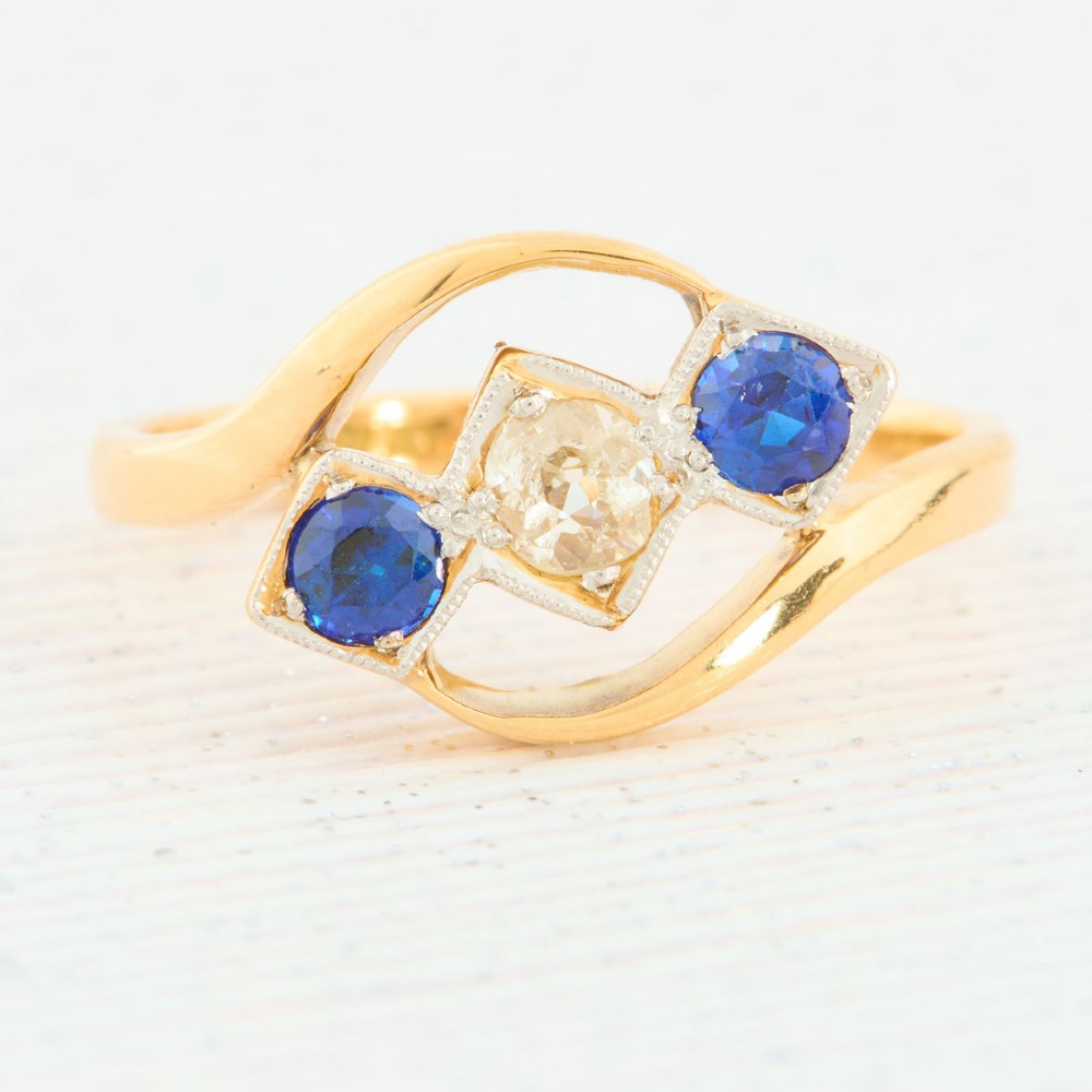 An Art Deco 18ct yellow gold dress ring featuring a central round diamond flanked by two round blue sapphires set diagonally in square millegrain mounts within a bypass band.