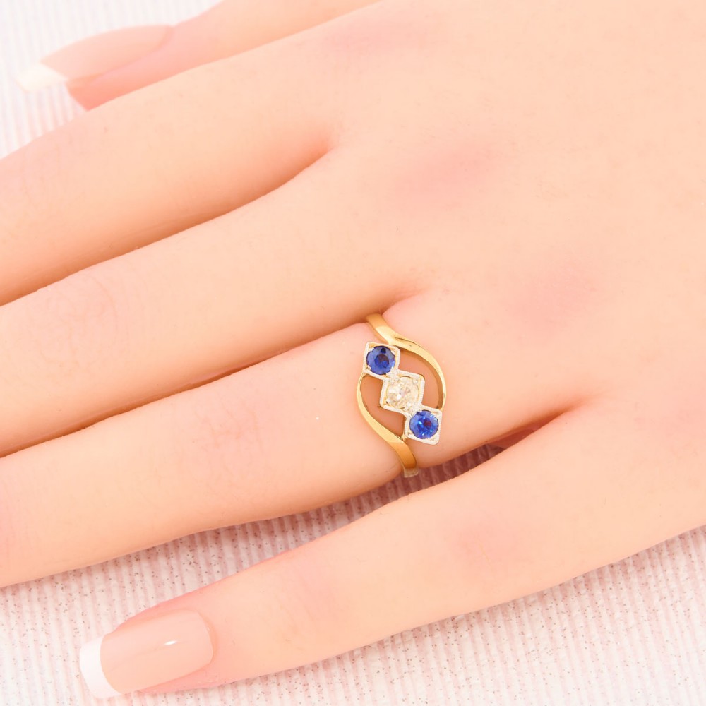 An Art Deco 18ct yellow gold dress ring featuring a central round diamond flanked by two round blue sapphires set diagonally in square millegrain mounts within a bypass band. View II