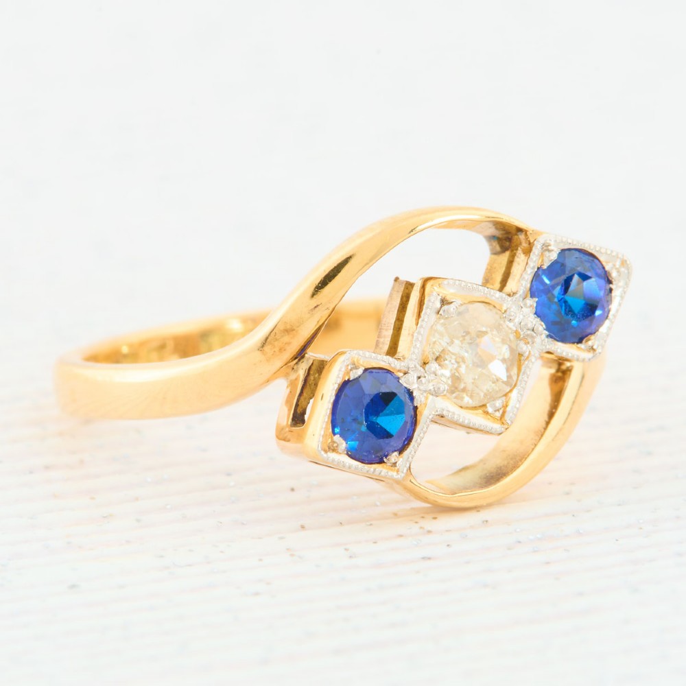An Art Deco 18ct yellow gold dress ring featuring a central round diamond flanked by two round blue sapphires set diagonally in square millegrain mounts within a bypass band. View III