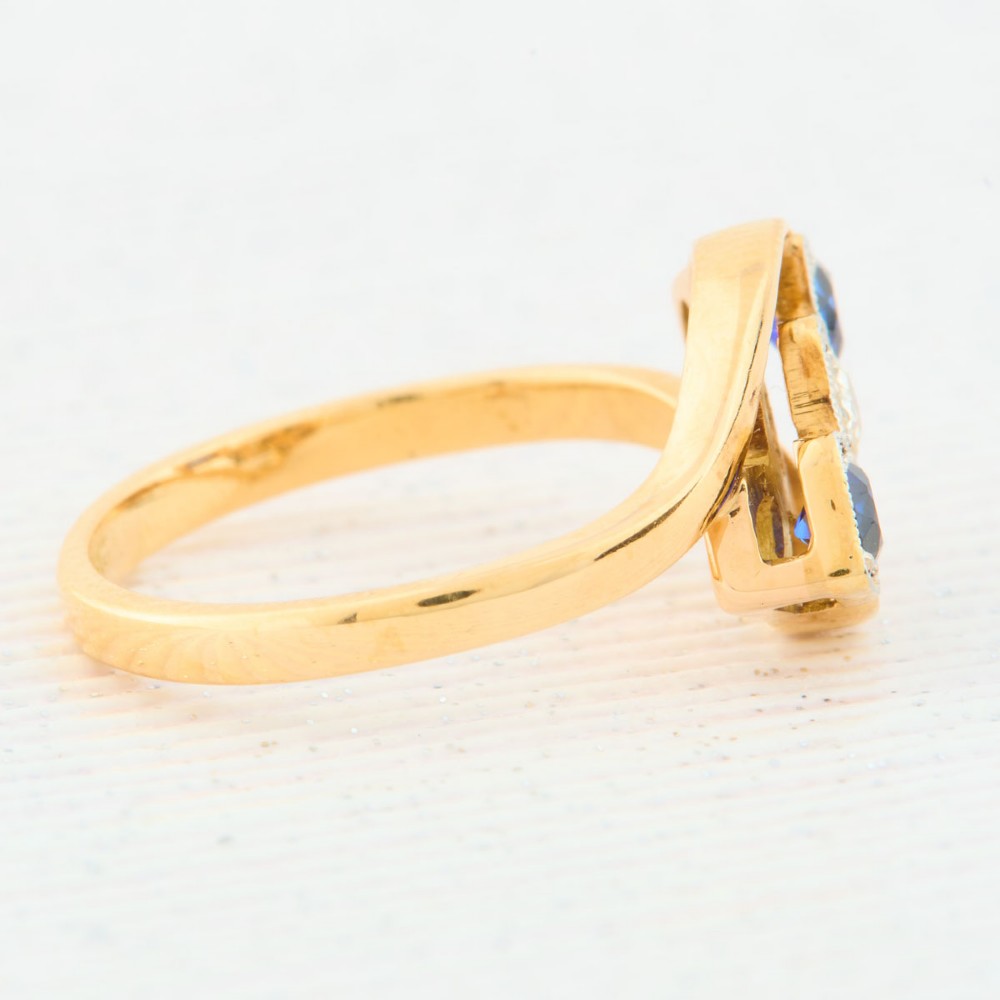 An Art Deco 18ct yellow gold dress ring featuring a central round diamond flanked by two round blue sapphires set diagonally in square millegrain mounts within a bypass band. View IV