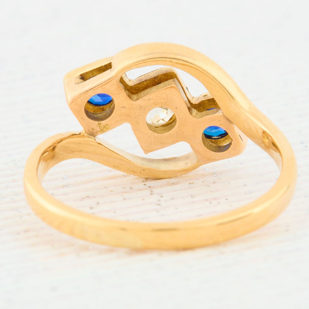 An Art Deco 18ct yellow gold dress ring featuring a central round diamond flanked by two round blue sapphires set diagonally in square millegrain mounts within a bypass band. View V