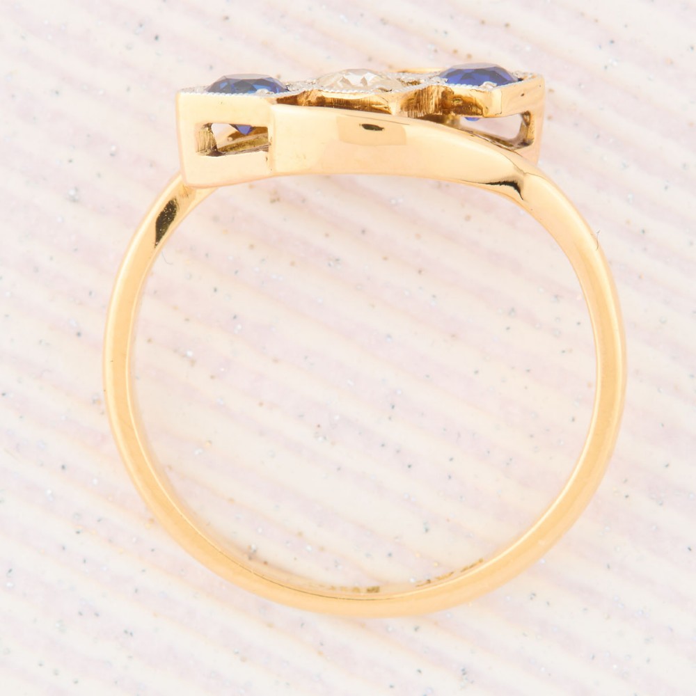 An Art Deco 18ct yellow gold dress ring featuring a central round diamond flanked by two round blue sapphires set diagonally in square millegrain mounts within a bypass band. View VI