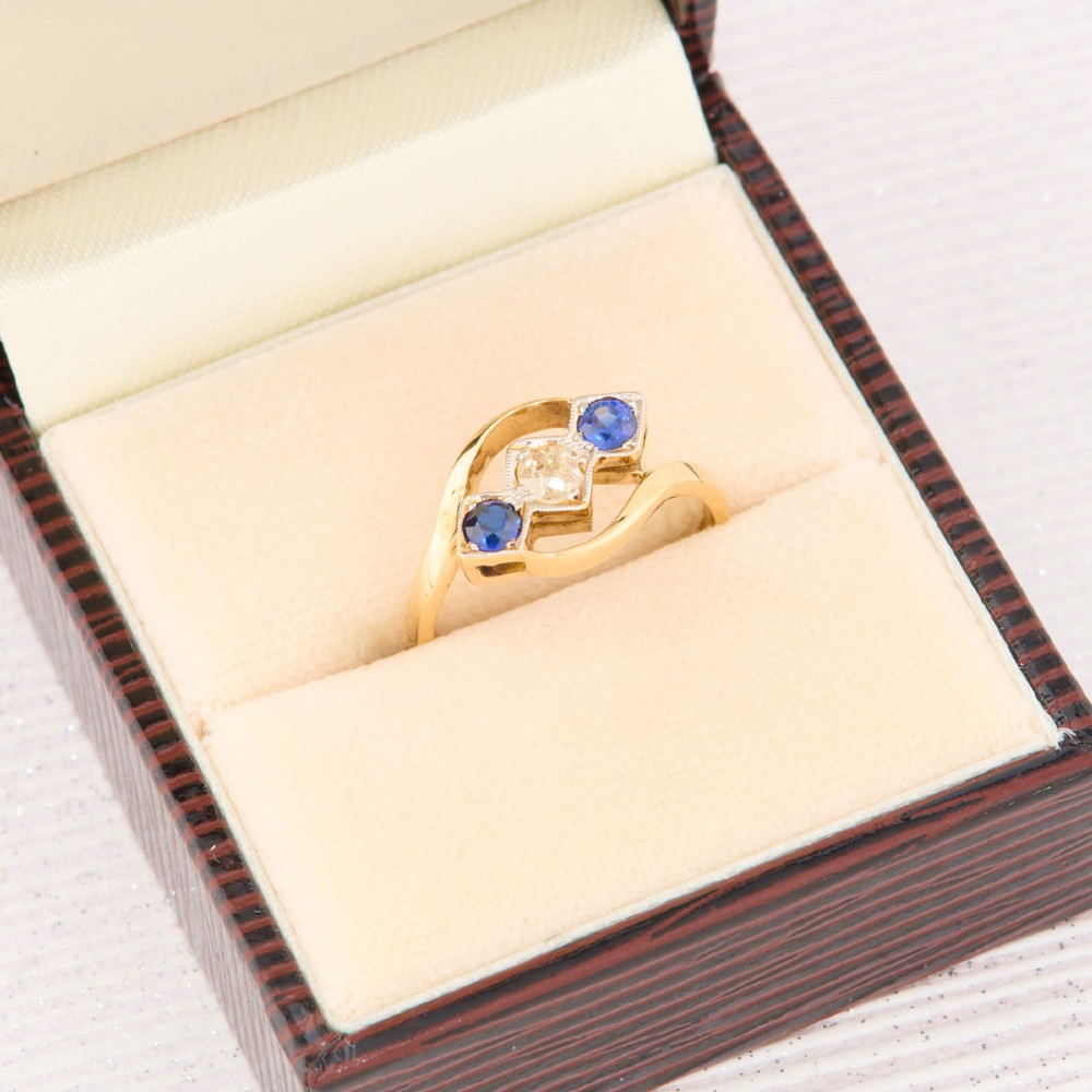 An Art Deco 18ct yellow gold dress ring featuring a central round diamond flanked by two round blue sapphires set diagonally in square millegrain mounts within a bypass band. View VIII
