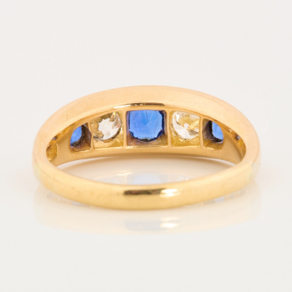An 18ct yellow gold five-stone ring featuring three round blue sapphires alternating with two round diamonds in a carved boat-style gallery. View V