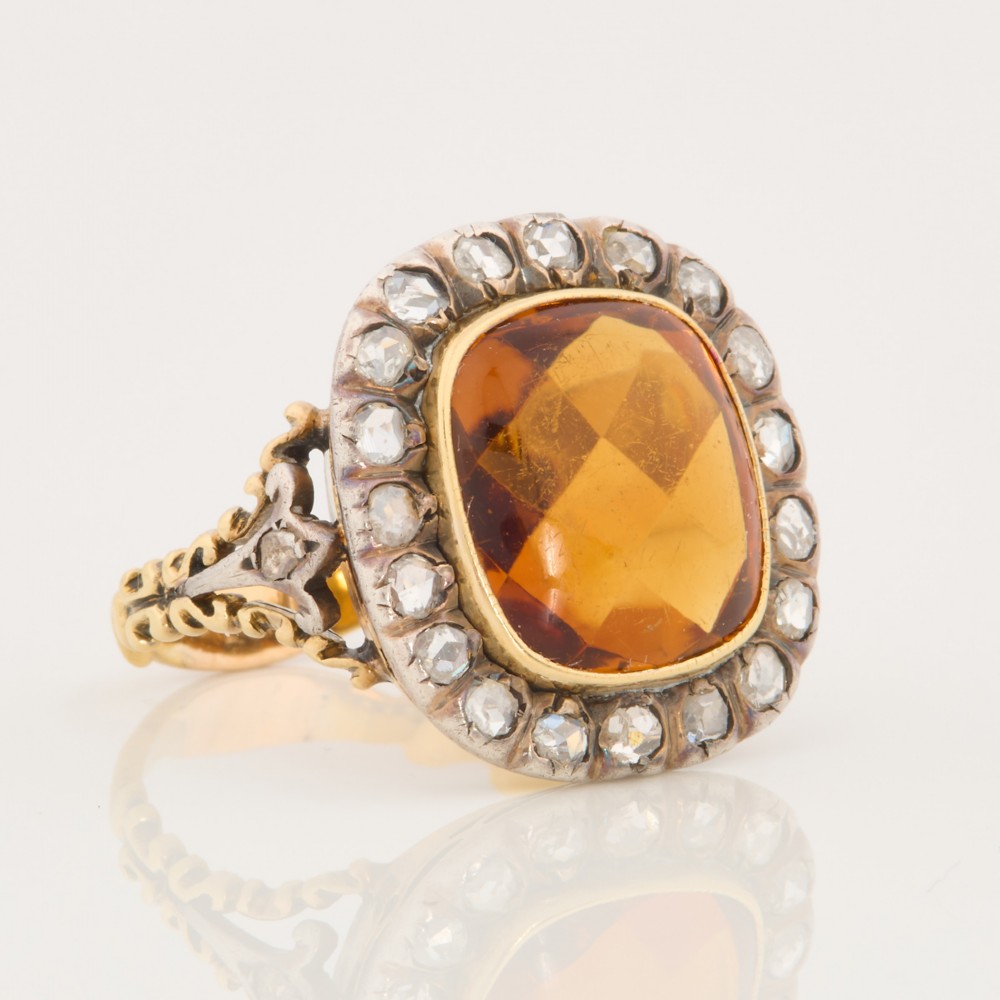An antique 18ct gold ring set with a central cushion-cut amber citrine encircled by a halo of diamonds with decorative scrollwork shoulders.