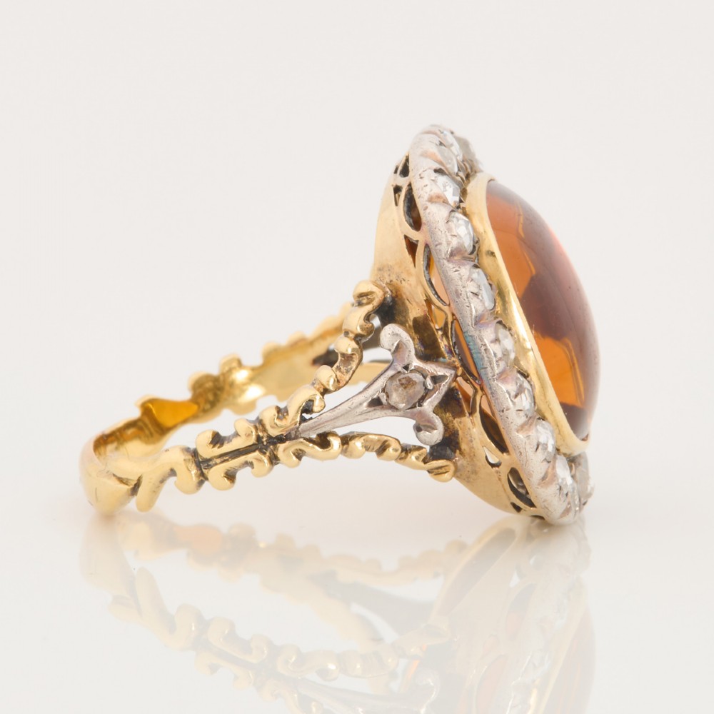 An antique 18ct gold ring set with a central cushion-cut amber citrine encircled by a halo of diamonds with decorative scrollwork shoulders. View IV