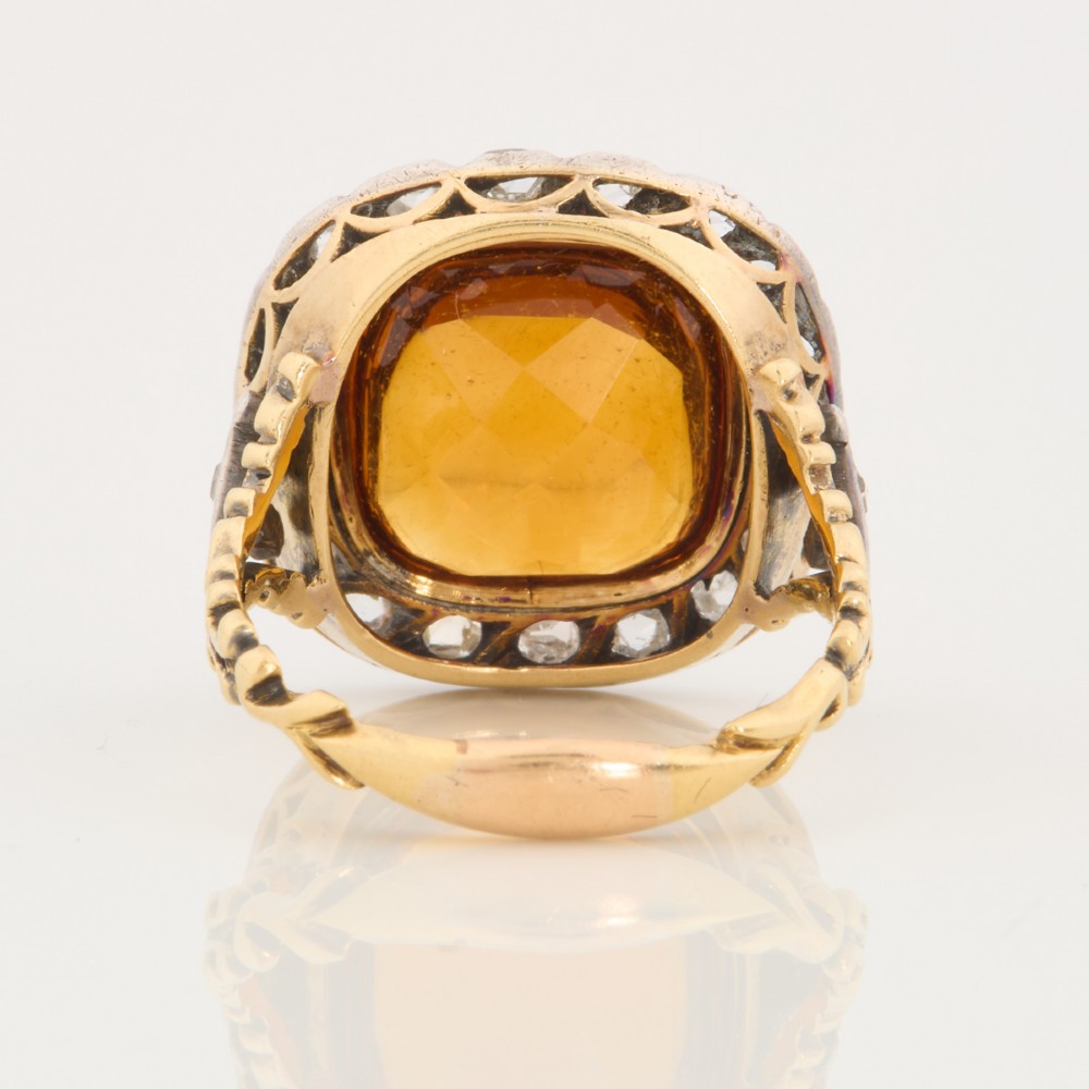 An antique 18ct gold ring set with a central cushion-cut amber citrine encircled by a halo of diamonds with decorative scrollwork shoulders. View V