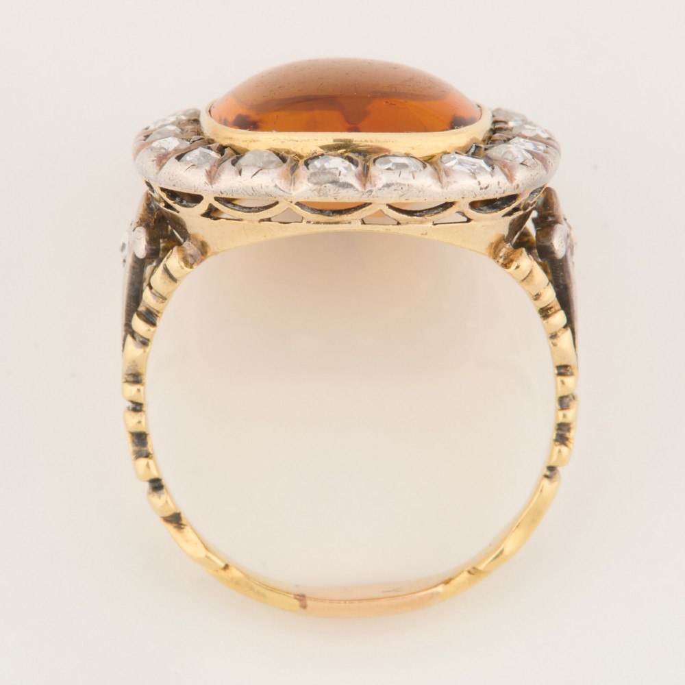 An antique 18ct gold ring set with a central cushion-cut amber citrine encircled by a halo of diamonds with decorative scrollwork shoulders. View VI