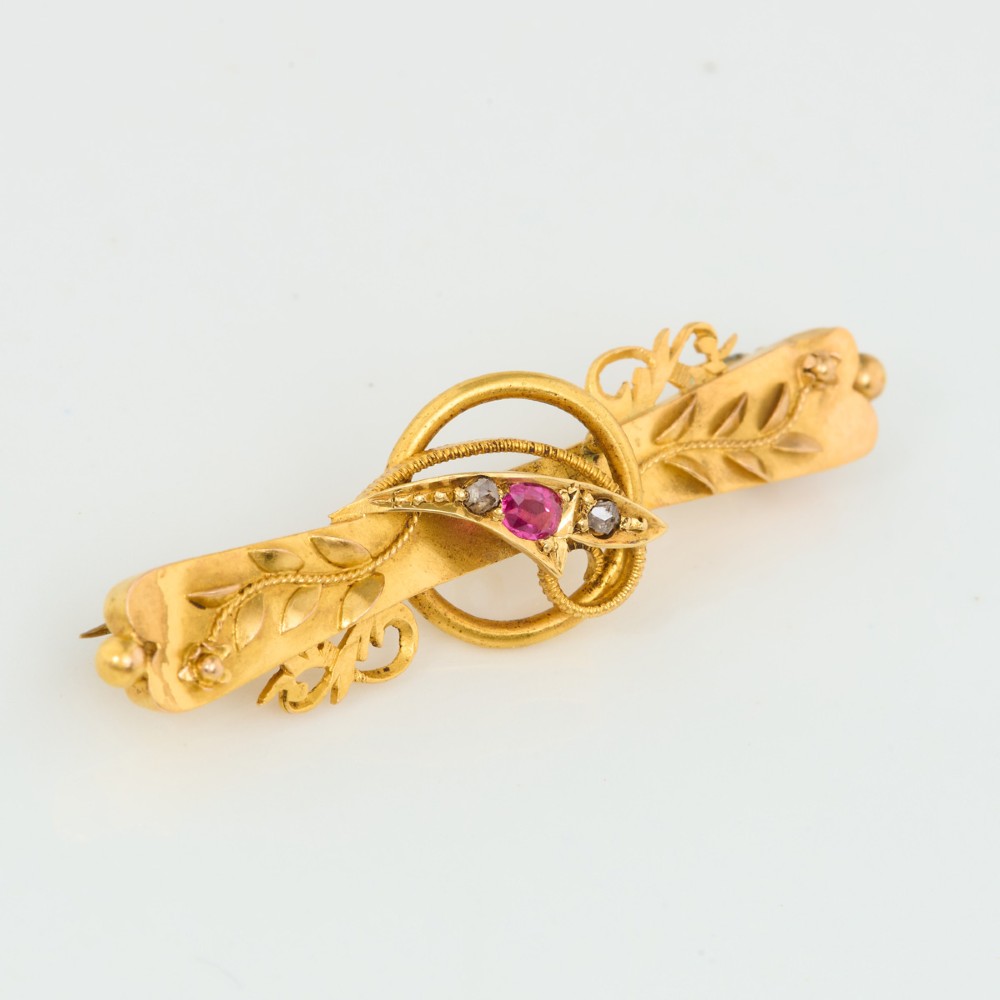 A yellow gold bar brooch featuring carved leaf detail, a central circular wire ring, and a central ruby flanked by two diamonds.