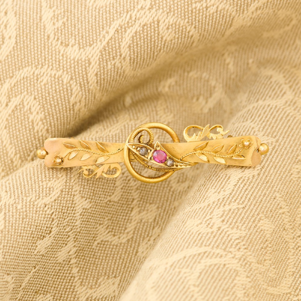 A yellow gold bar brooch featuring carved leaf detail, a central circular wire ring, and a central ruby flanked by two diamonds. View II
