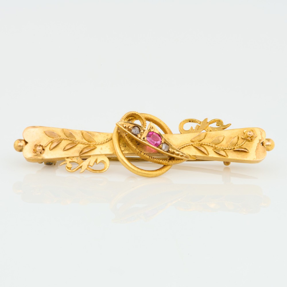 A yellow gold bar brooch featuring carved leaf detail, a central circular wire ring, and a central ruby flanked by two diamonds. View III