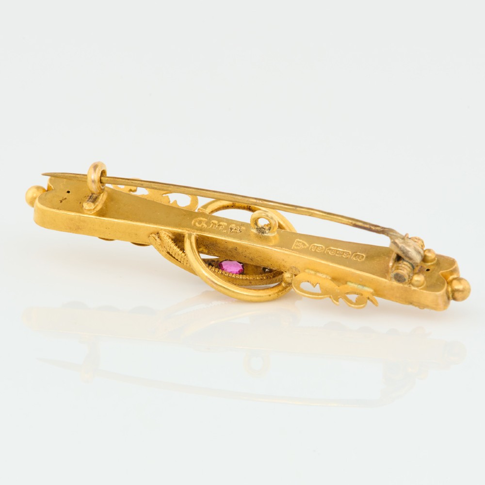 A yellow gold bar brooch featuring carved leaf detail, a central circular wire ring, and a central ruby flanked by two diamonds. View V