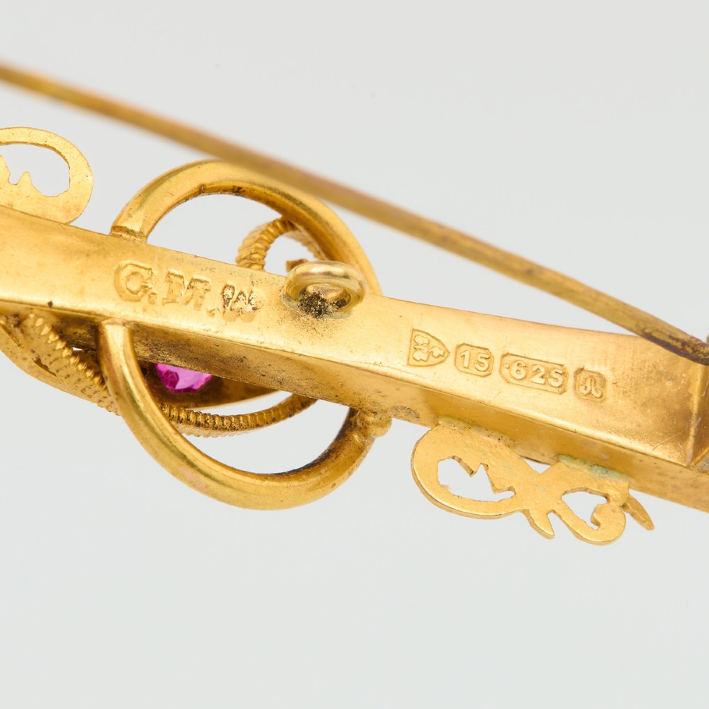 A yellow gold bar brooch featuring carved leaf detail, a central circular wire ring, and a central ruby flanked by two diamonds. View VI