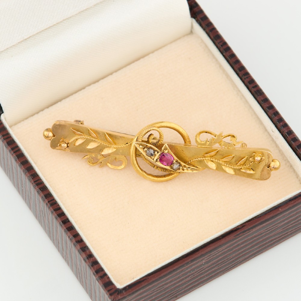 A yellow gold bar brooch featuring carved leaf detail, a central circular wire ring, and a central ruby flanked by two diamonds. View VII