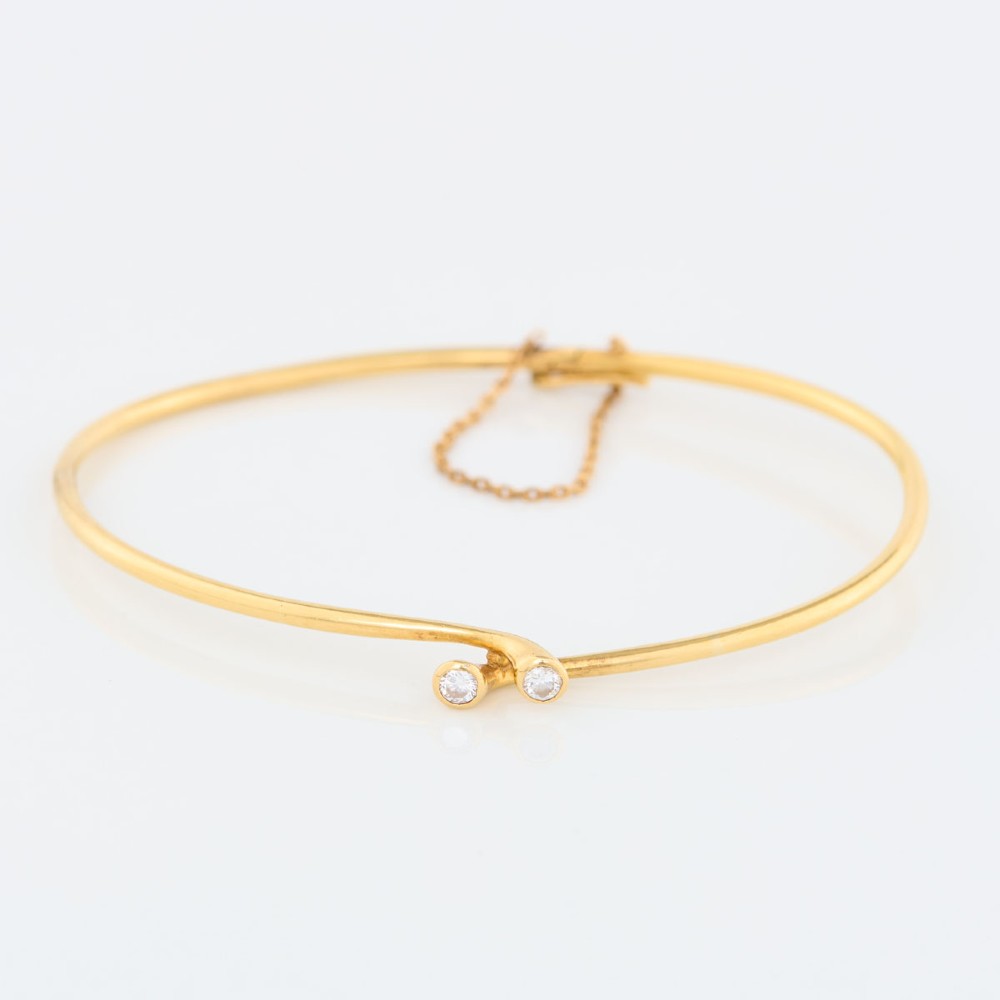 A slim 18ct yellow gold crossover bangle featuring two bezel-set diamonds at the open ends and an attached safety chain.