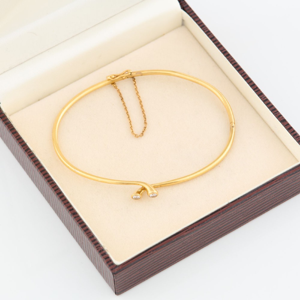 A slim 18ct yellow gold crossover bangle featuring two bezel-set diamonds at the open ends and an attached safety chain. View VI