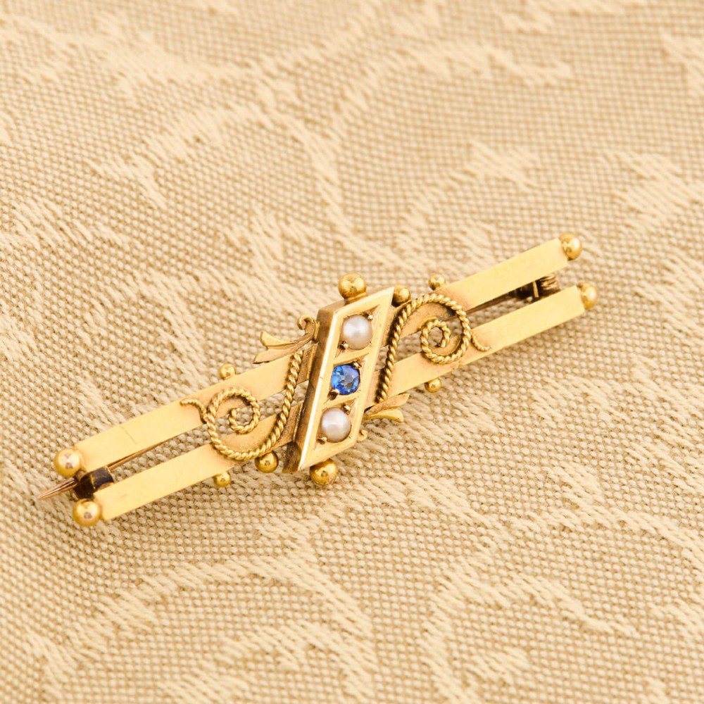 A late Victorian 15ct gold double bar brooch featuring a central lozenge setting with a round blue sapphire flanked by two pearls, decorated with rope-twist wirework and ball finials. View II
