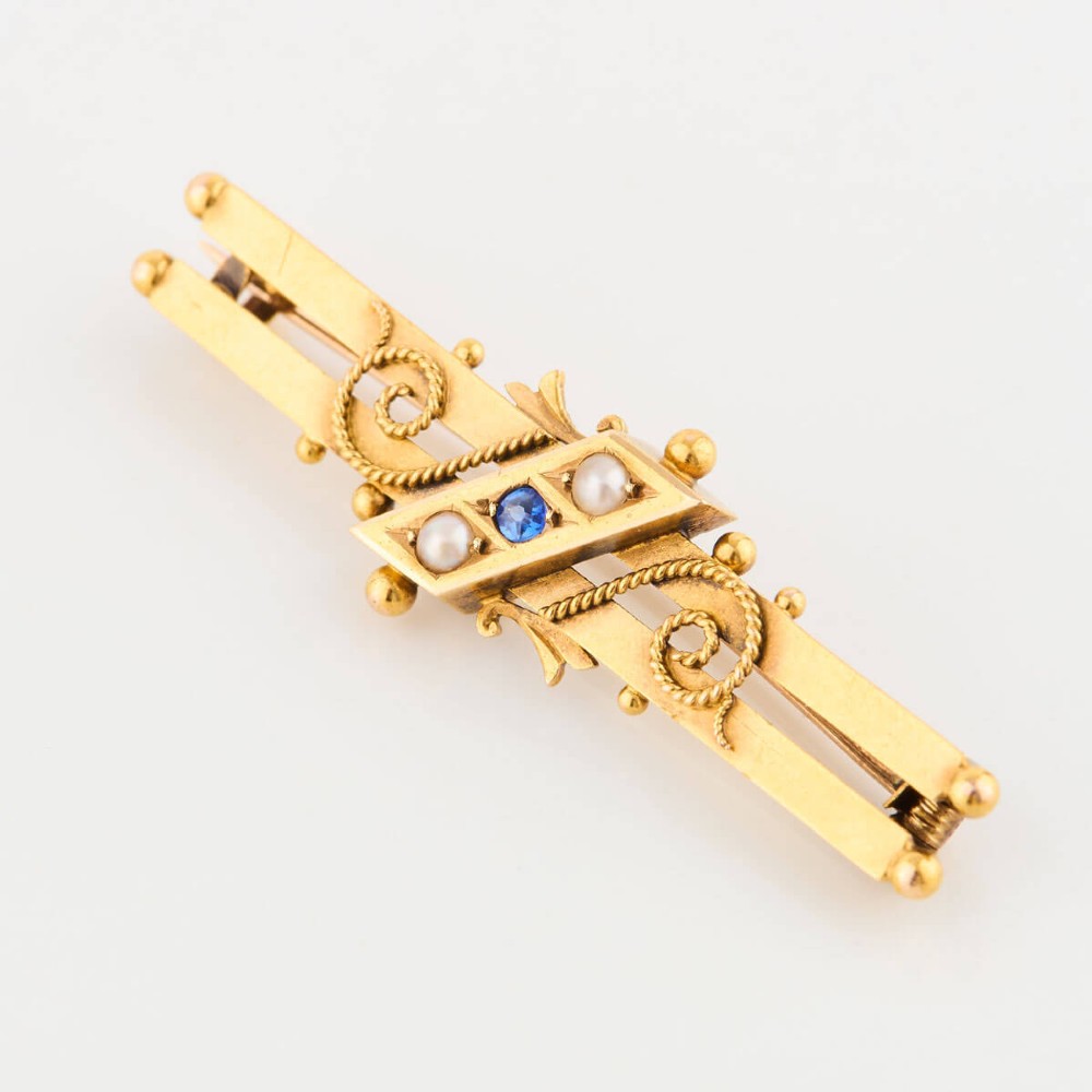 A late Victorian 15ct gold double bar brooch featuring a central lozenge setting with a round blue sapphire flanked by two pearls, decorated with rope-twist wirework and ball finials. View III