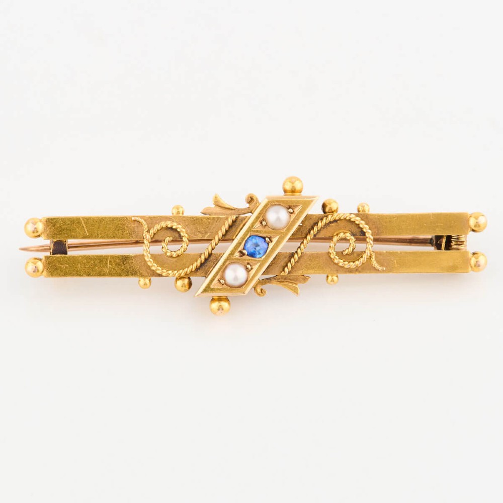A late Victorian 15ct gold double bar brooch featuring a central lozenge setting with a round blue sapphire flanked by two pearls, decorated with rope-twist wirework and ball finials. View IV