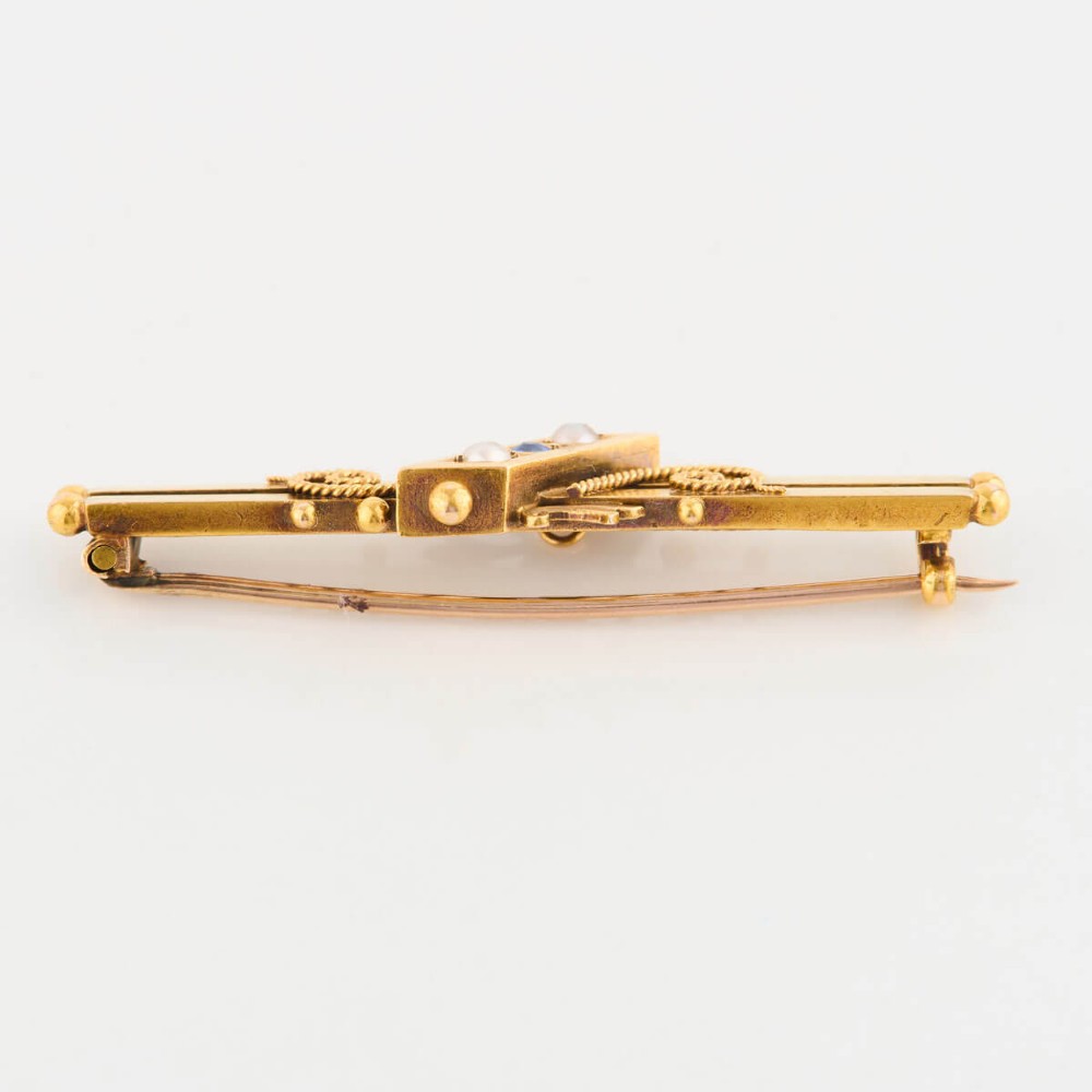 A late Victorian 15ct gold double bar brooch featuring a central lozenge setting with a round blue sapphire flanked by two pearls, decorated with rope-twist wirework and ball finials. View V