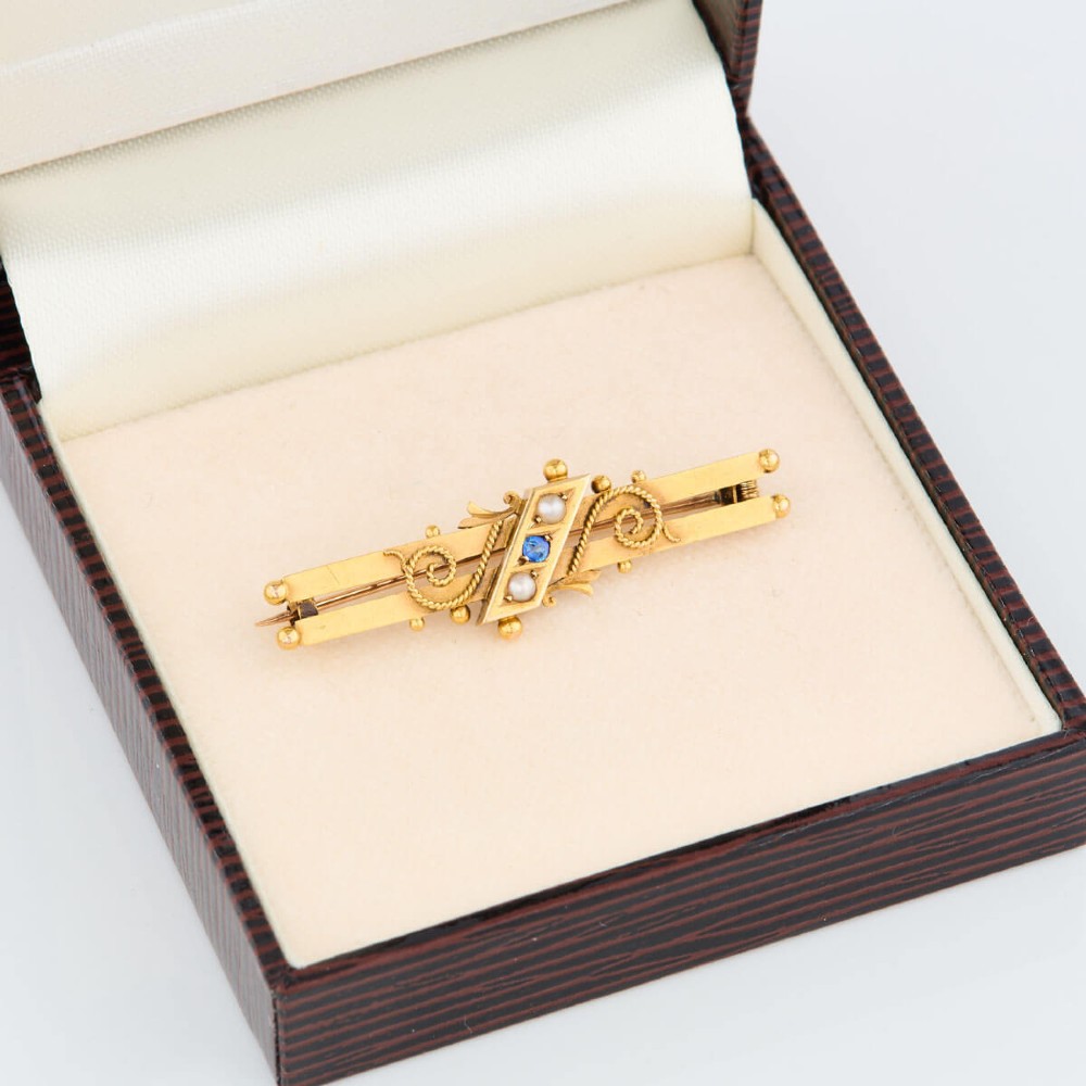 A late Victorian 15ct gold double bar brooch featuring a central lozenge setting with a round blue sapphire flanked by two pearls, decorated with rope-twist wirework and ball finials. View VIII