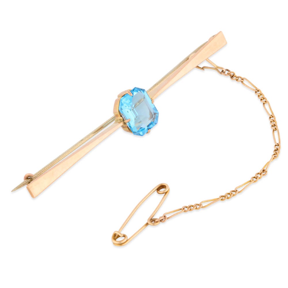 A 9ct yellow gold horizontal bar brooch with a central emerald-cut blue paste stone and an attached safety chain.