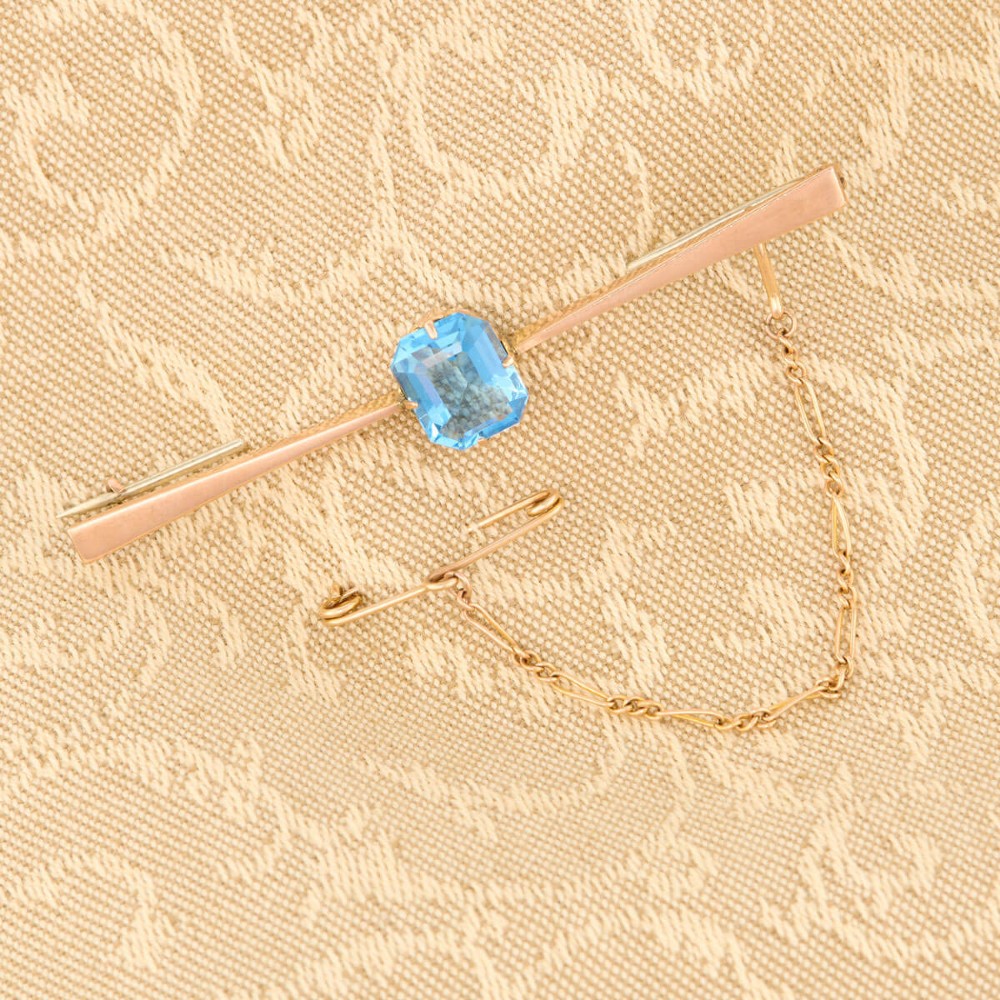A 9ct yellow gold horizontal bar brooch with a central emerald-cut blue paste stone and an attached safety chain. View II