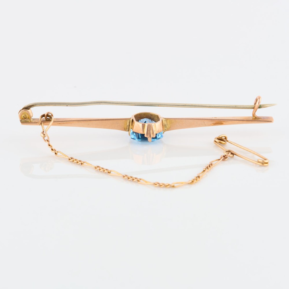 A 9ct yellow gold horizontal bar brooch with a central emerald-cut blue paste stone and an attached safety chain. View IV