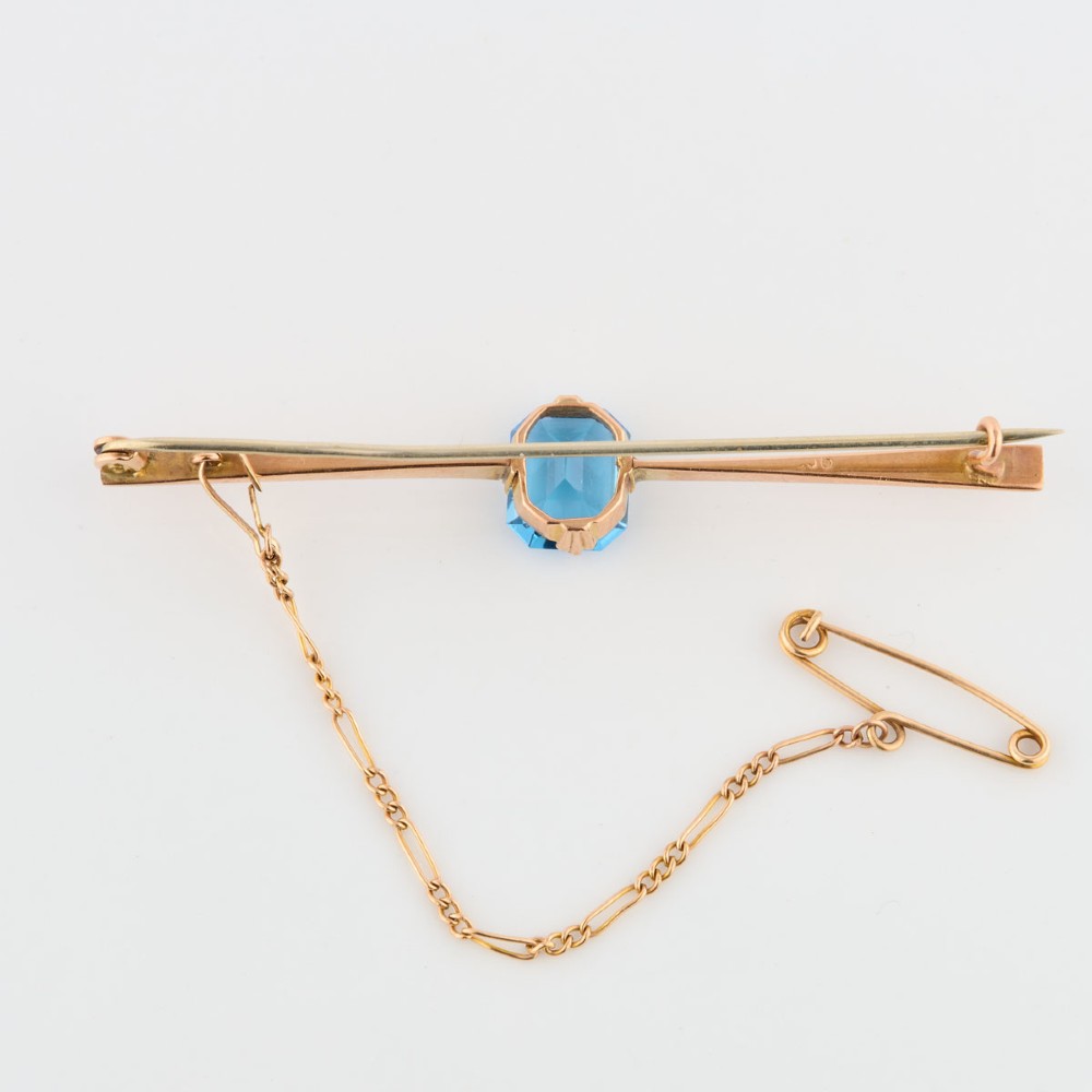 A 9ct yellow gold horizontal bar brooch with a central emerald-cut blue paste stone and an attached safety chain. View V